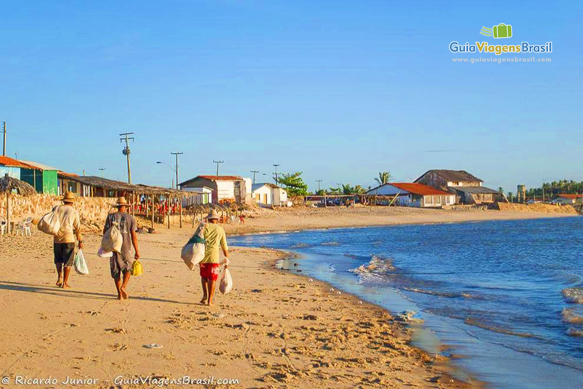 Imagem de moradores carregando sacolas pela praia. Faixa de areia pequena com casa simples pela orla.